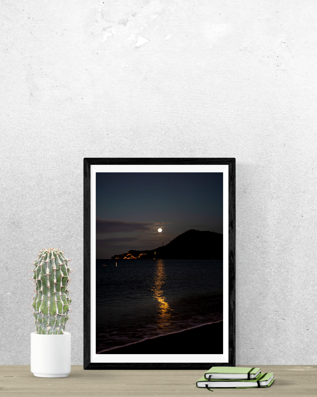 Framed artwork of a moonlit landscape over water on a gray wall with a cactus and folded blanket on a table.